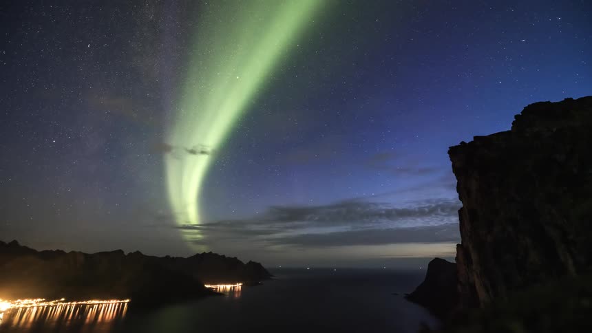 The green lights of the aurora borealis move among the stars in the night sky as lights reflect on the water