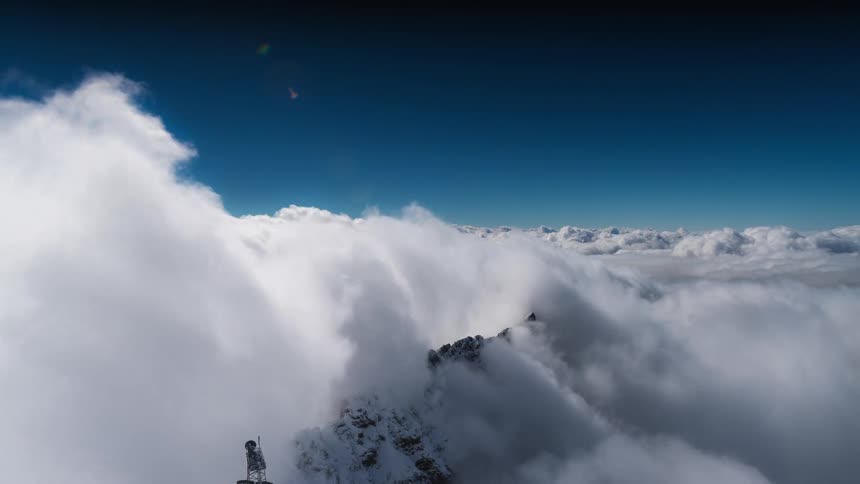 View from above the clouds as they move over a mountain with a statue on top in a clear blue sky