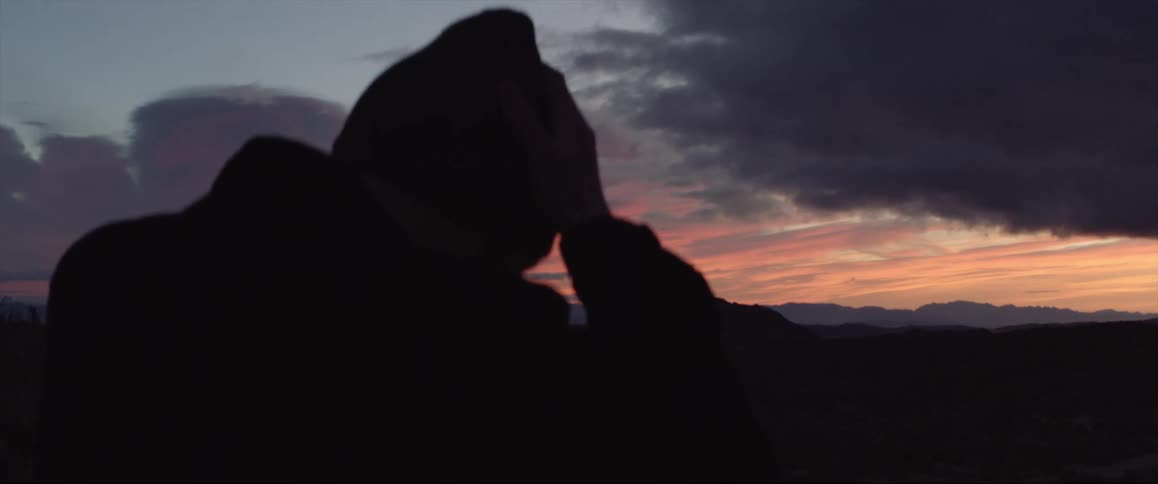 A man puts on a beanie while sitting outside as the sun sets