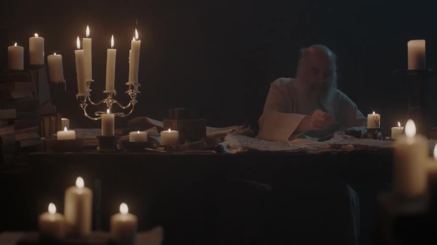 A bald man with a long white beard sits at a table and writes by candle light