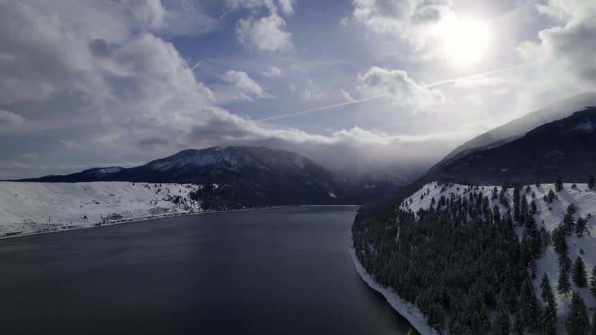 Conifer trees cover snowy slopes bordering water under clouds