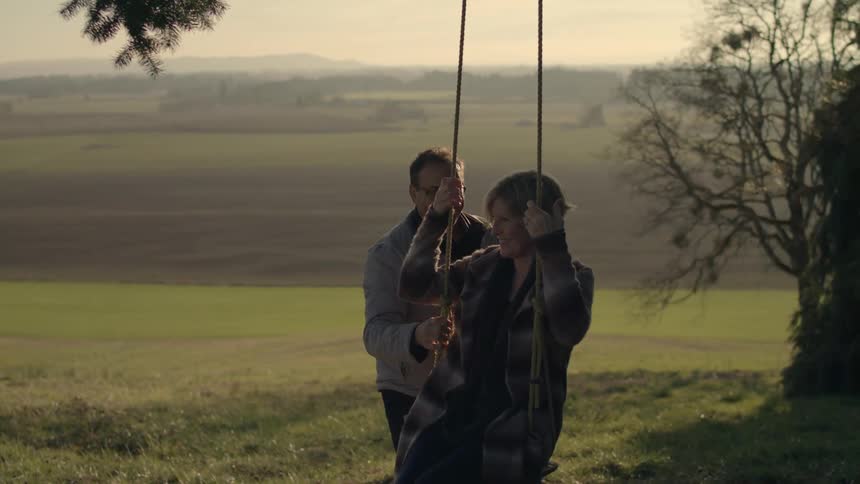 Mature couple play in a swing under a tree in the countryside at evening
