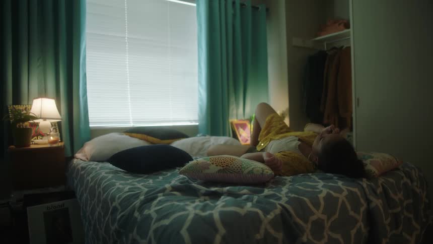 Woman wearing white blouse and yellow shirt lies on bed talking on smartphone in her room