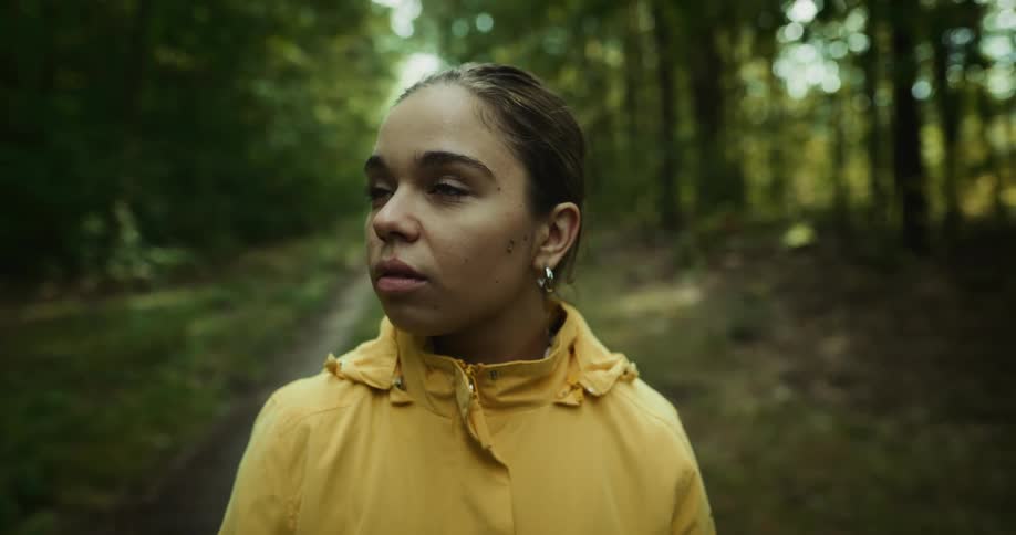 Woman with bun earrings and yellow raincoat looks up at trees in the green woods