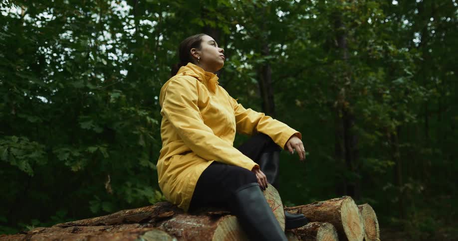 A woman in a yellow raincoat sits atop a pile of logs while looking into the distance