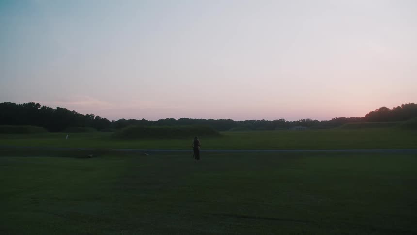 A woman walks across an open field at sunset