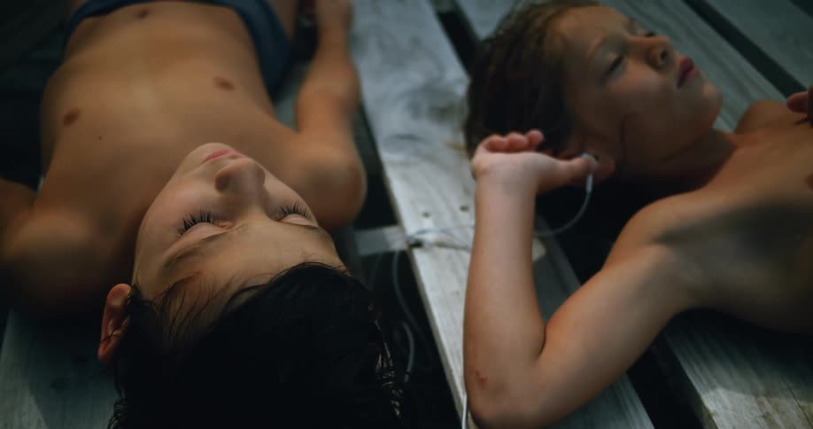 Two boys wearing shorts are lying on top of a picnic table listening to earphones together as each boy has one in his ear
