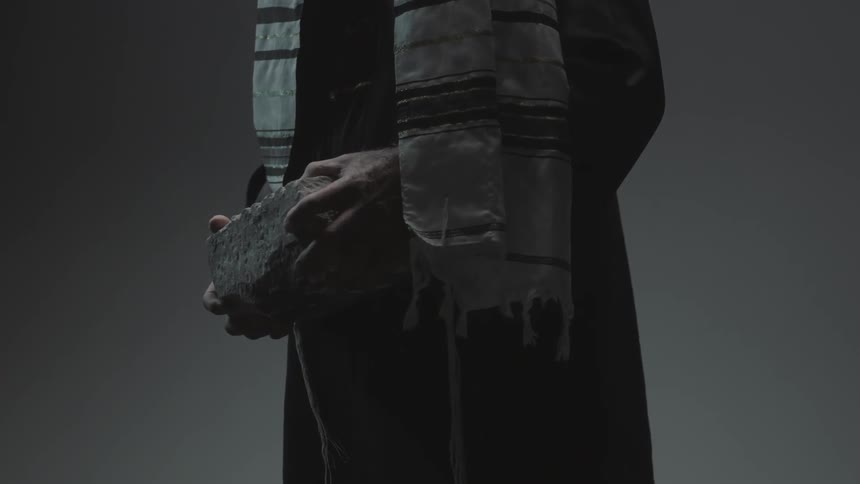 A man wearing a scarf stands in a dim room while holding a large rock