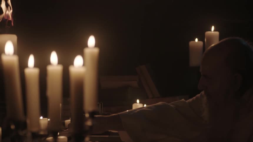 A bald man with a long white beard sits at a table and writes by candle light