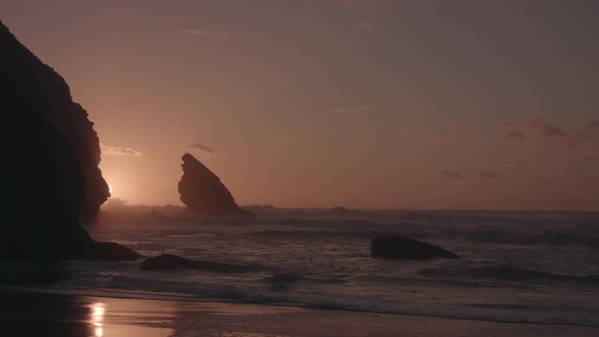 A pink sunrisesunset on the horizon with waves crashing against the coastline with a silhouette of a rock formation in the water