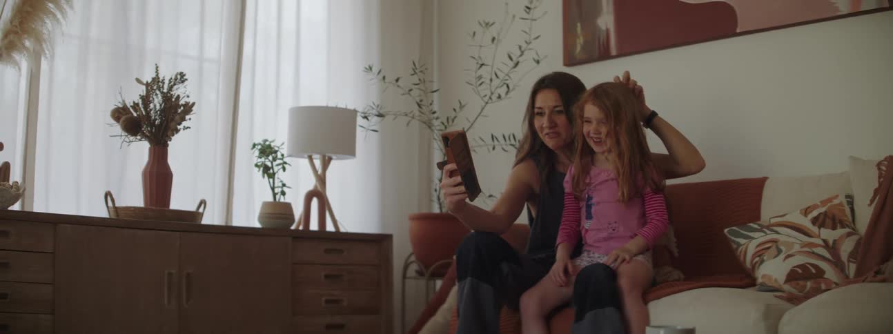 Mother with brown hair gray top and pants holds daughter with red head kissing and taking a selfie with smartphone in living room at home