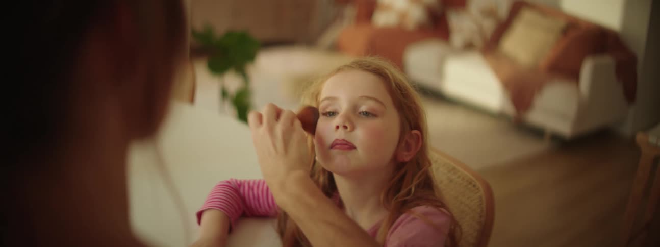 A little girl is sitting in a chair at a table as her mother brushes make up on her face