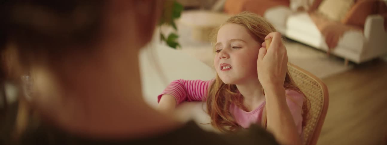 A mother sits at home with her young daughter and brushes a make up brush over her face