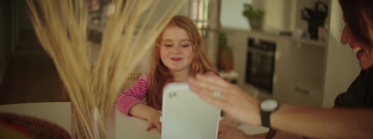 A little girl is sitting at a table with her mother and she smiles when her mother holds up a toy for her to see