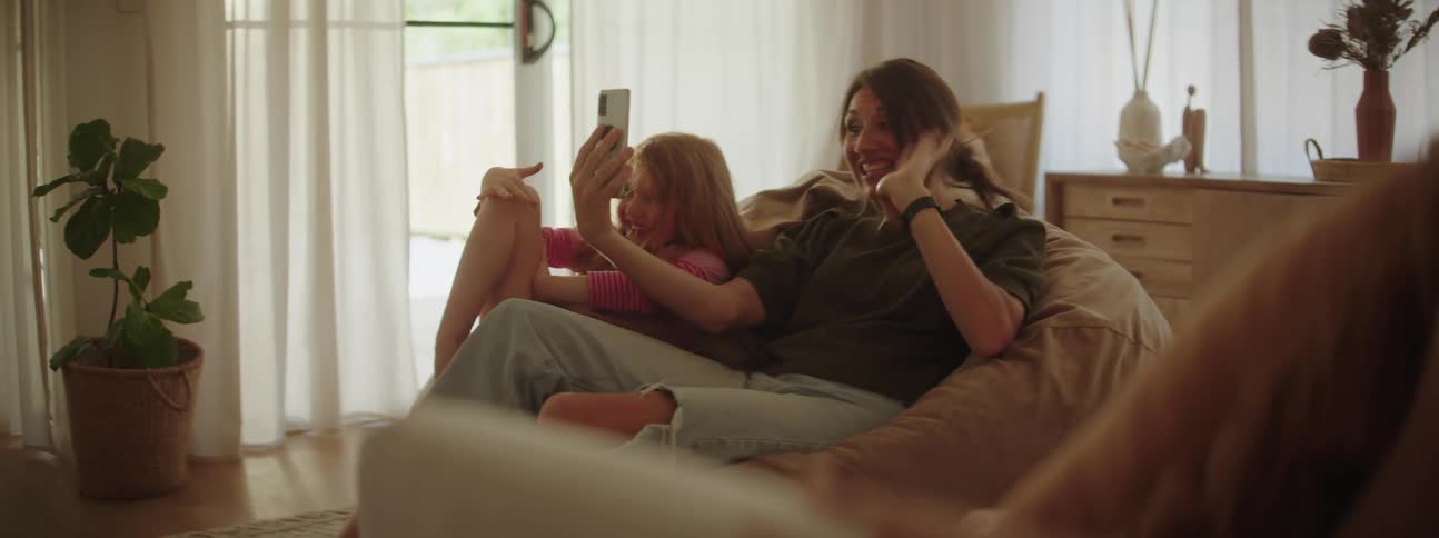 Mother with ponytail gray t shirt and ripped jeans and daughter with red hair make video call on smartphone waving in living room at home