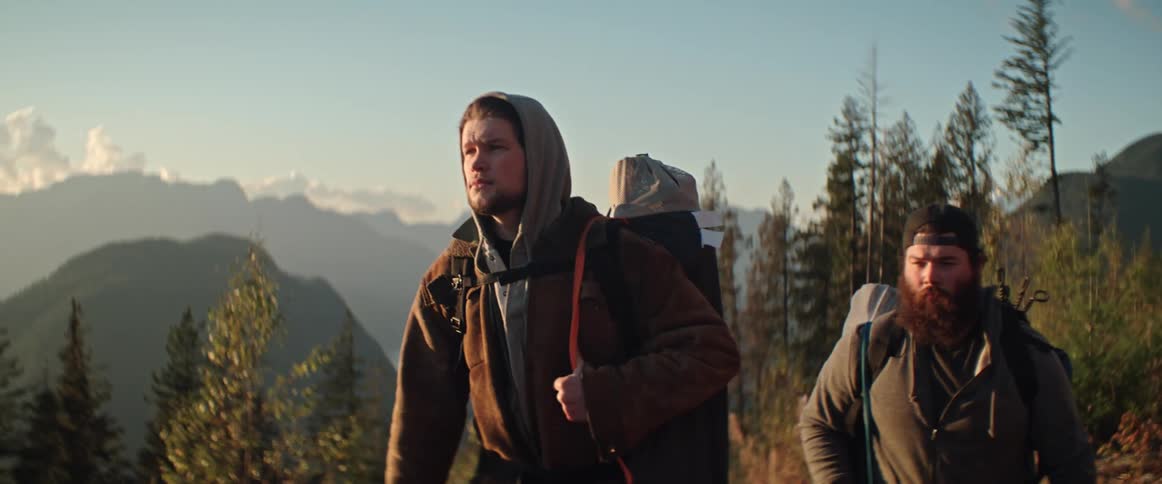 Two men backpack through a forest clearing under a mountain range