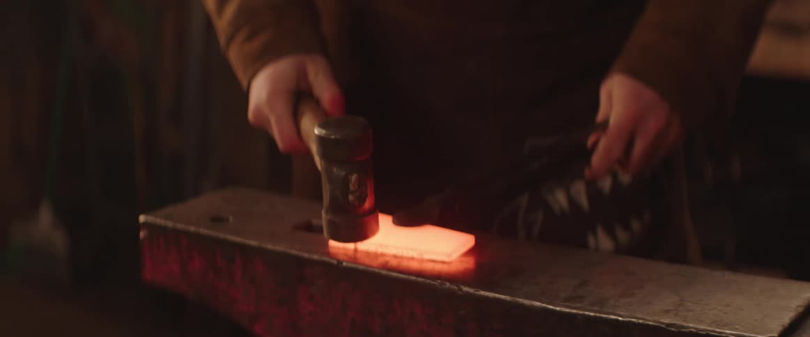 Blacksmith hammers molten piece of metal being held with pincers on anvil