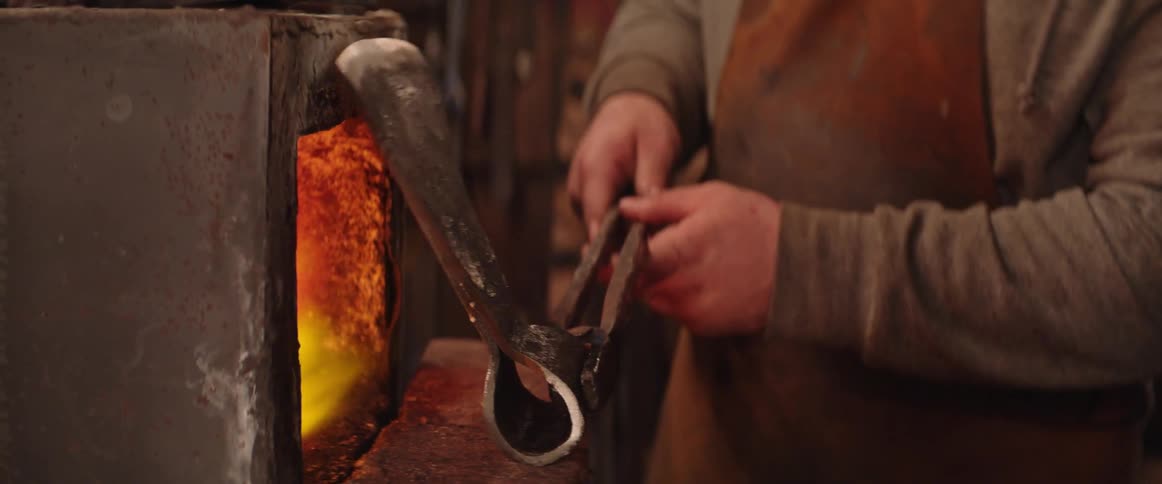Blacksmith holding metal axe head with tongs heats the object at forge