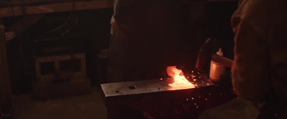 Two blacksmiths forge an axe head by alternating hammer swings onto the hot metal