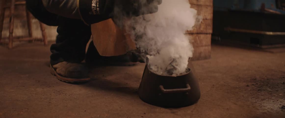 A plume of smoke rises after a blacksmith man places a hot axe head in a pot of oil