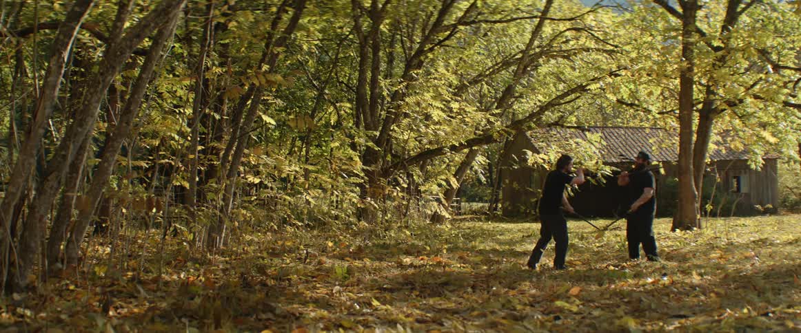 Two men practice fighting with sticks in the backyard of a cabin in the woods