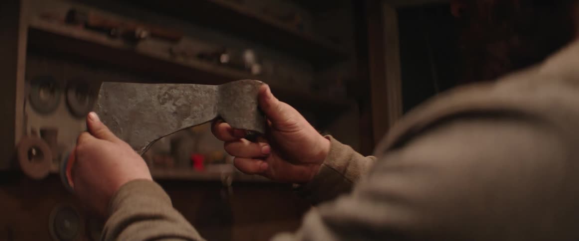 A craftsman is standing near a bench holding an axe blade and looking at it from many angles