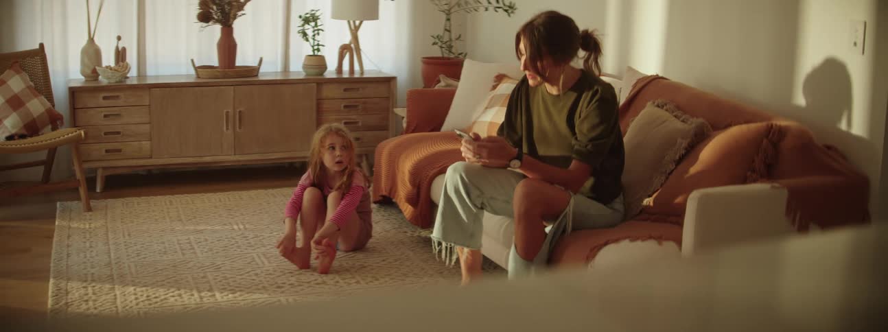 Mother with ponytail black t shirt and ripped jeans sits on couch using smartphone while talking to daughter sitting on rug in living room at home