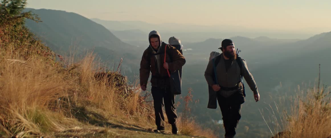 Two men wearing backpacks are hiking up a mountain out of a large valley