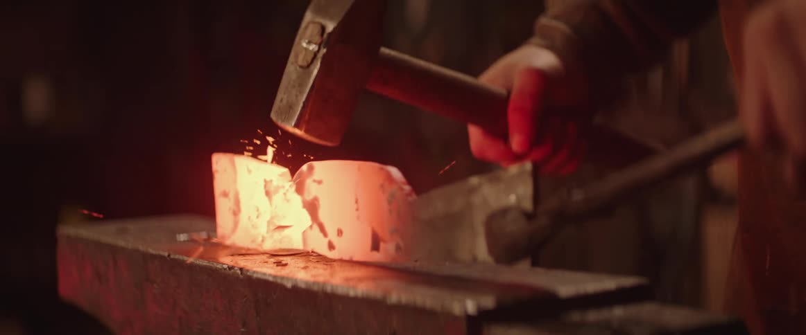 Blacksmith wearing apron forges an axe on an anvil as slag flies through the air