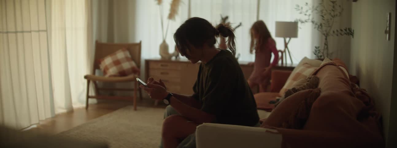 Woman with ponytail gray t shirt and ripped jeans sits on couch using smartphone in room while the girl standing in the corner