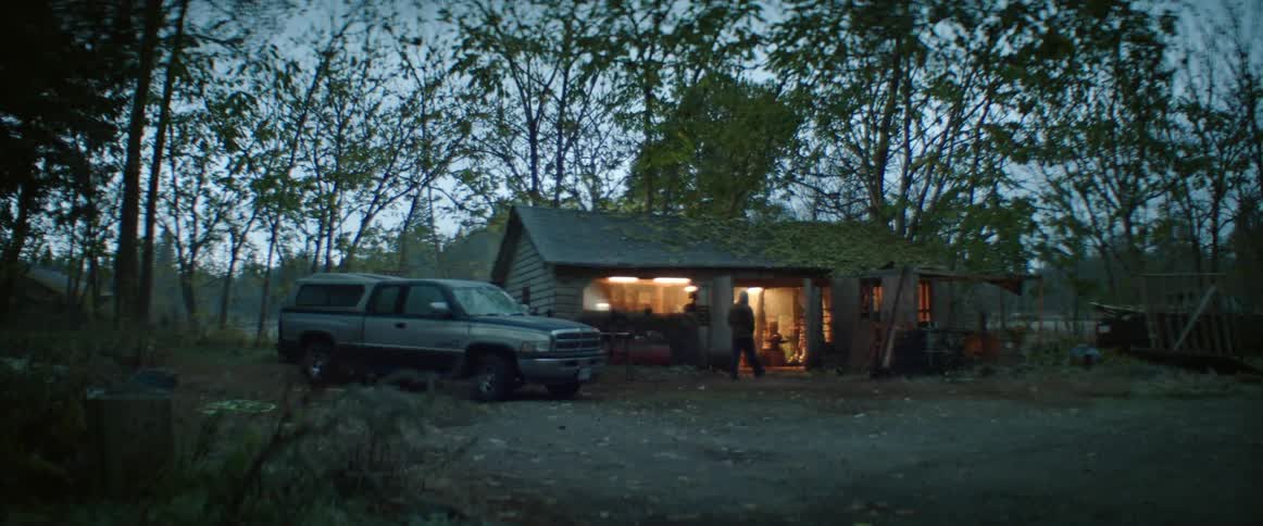 A person walks from their truck to a workshop with lights on in a rural area at dawn