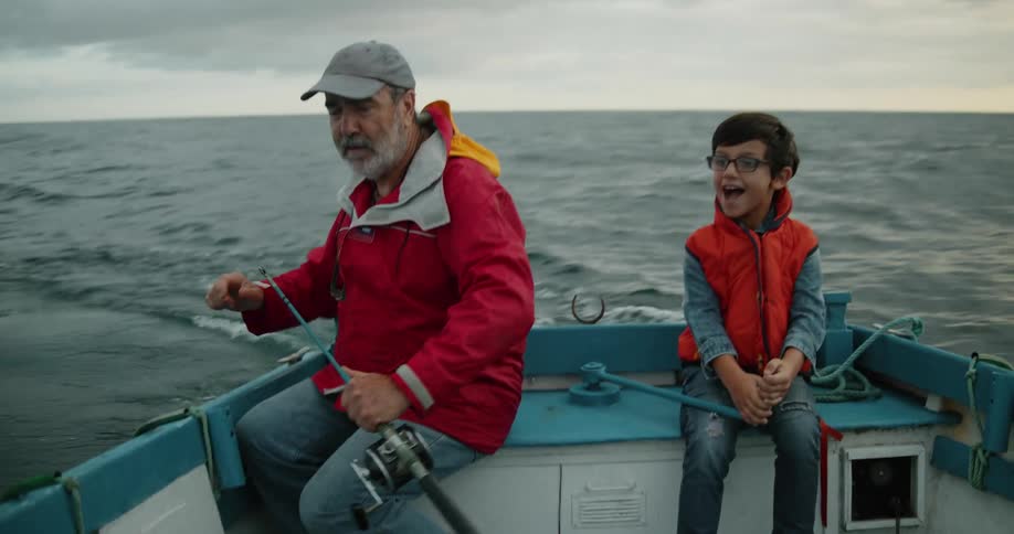 Young grandson sits in boat on water with grandfather as man spools out line from a fishing reel