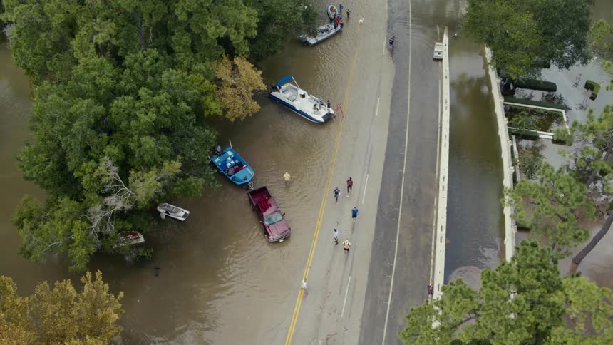 A small group of people walk past vehicles and boats along a flooded road
