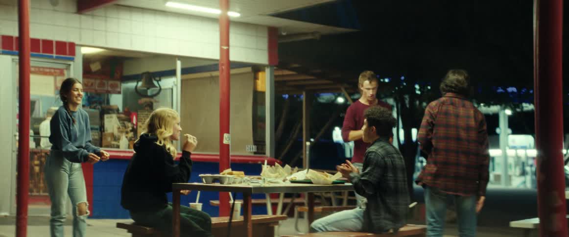 A group of teenage boys and girls sits at desk eating fast food and joking with each other