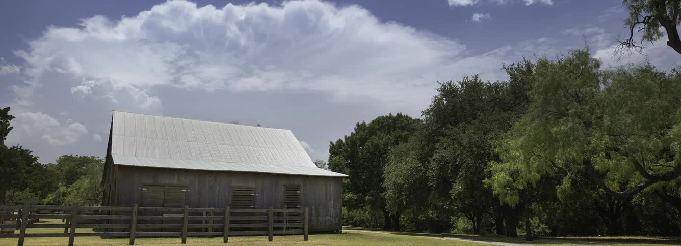 White clouds circle in the sky above a barn with an fence around it near a line of trees