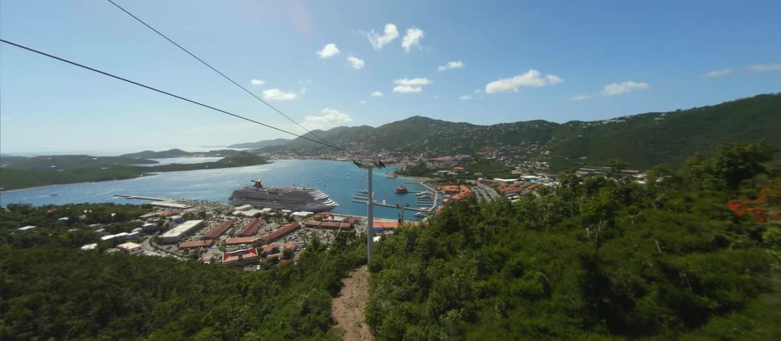 FPV - Aerial view of a green hillside with sky cars going down the rail and a cruise ship at the ocean port