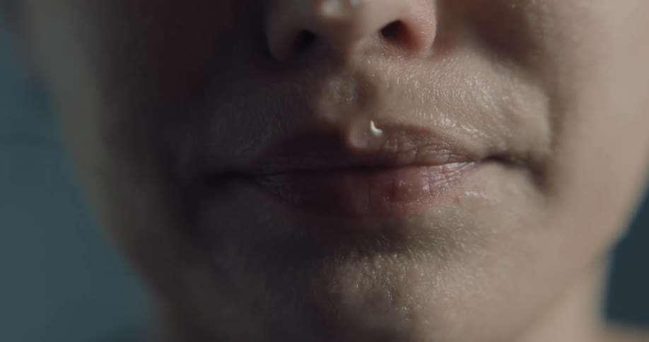 Lower face with droplet on upper lip is reflected in surface of clear liquid as drops fall and create ripples