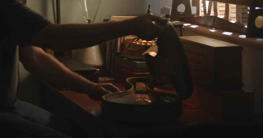A man sits at a desk in a home and opens a case to remove a small guitar in the daytime