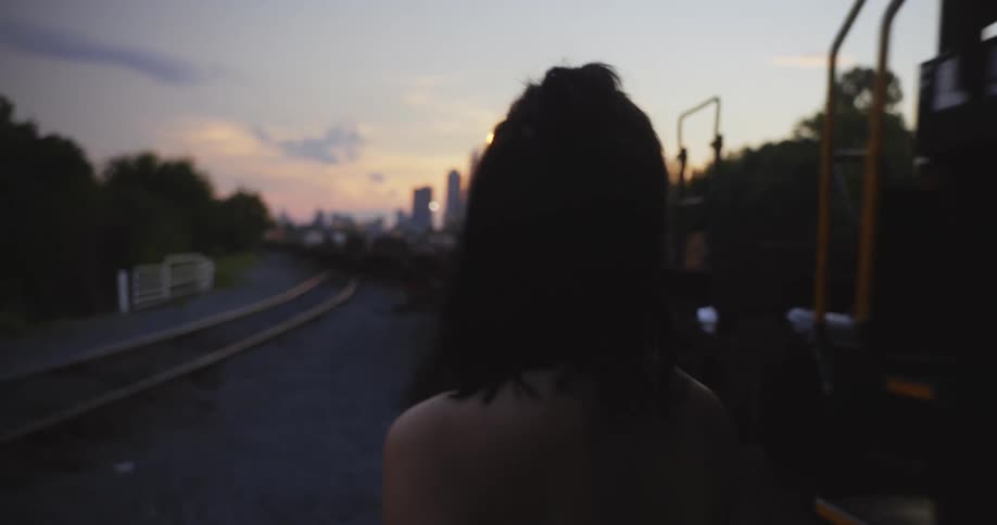 A young woman is standing in a train yard at dusk as a train passes by