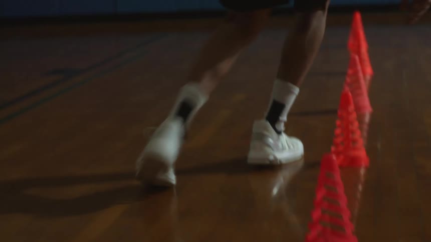A person wearing sneakers weaving in and out of cones dribbling a basketball