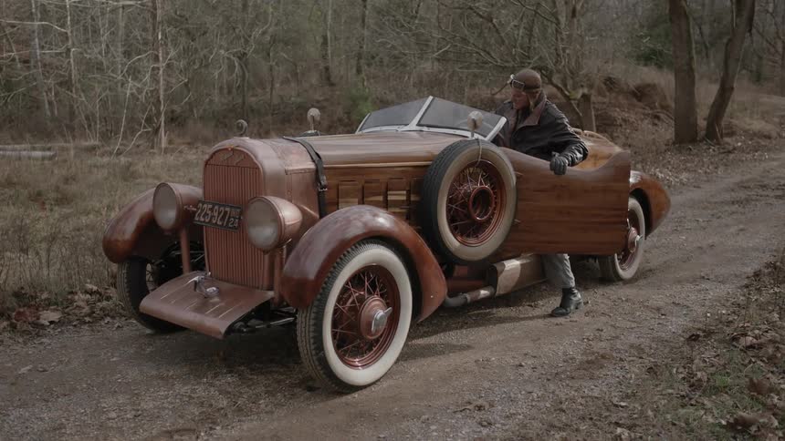 A man wearing a leather jacket and goggles enters an antique car near the woods in the daytime