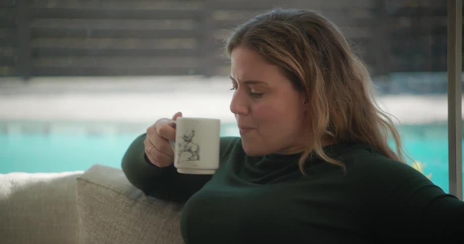 A woman lifts a coffee mug from a table and takes a sip while sitting on a sofa in the living room of a house during the day