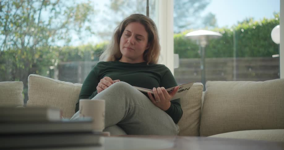 A woman reading a book on a sofa looks up while lost in thought