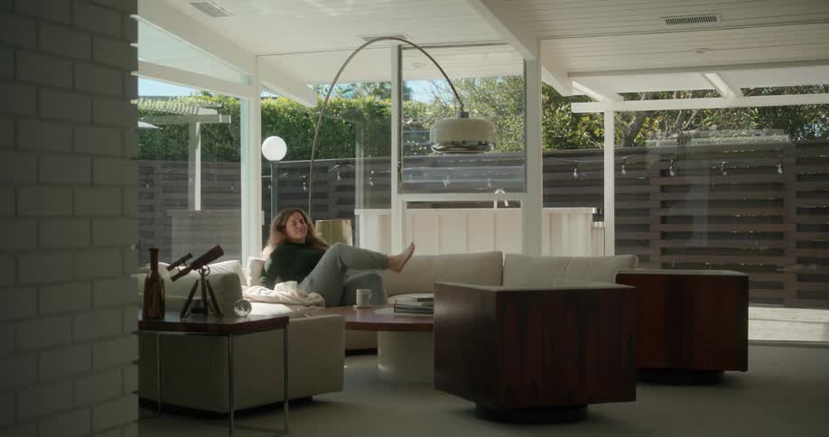 Woman carrying a cup of coffee and a coat on her arms sits down on the couch and starts stretching her arms and neck