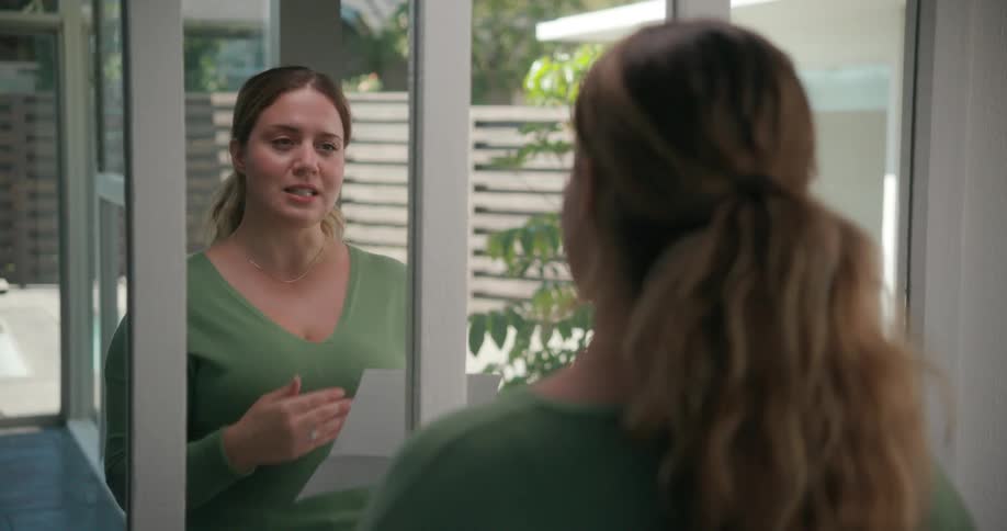 Woman with brown ponytail and green sweater looks in the mirror reading paper
