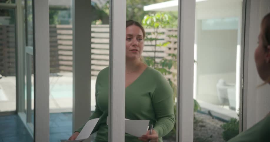 A woman is walking in front of a mirror speaking and looking at her notes on paper