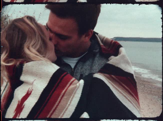 A man and woman standing on a beach wrapped up in a blanket and kissing