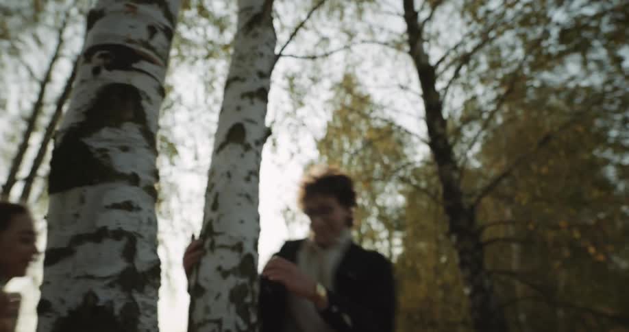 A teenage couple are running and playing chase around a tree near the woods