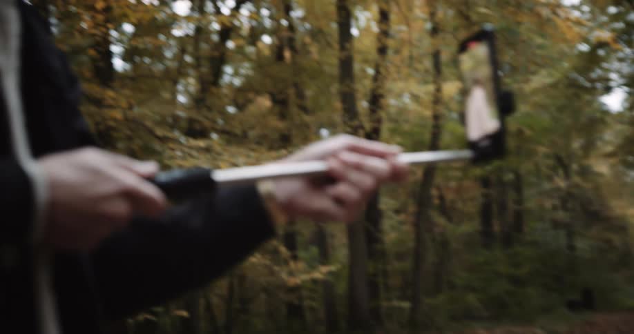Man with glasses black jacket and backpack films blonde woman wearing beige coat and gray scarf in the forest