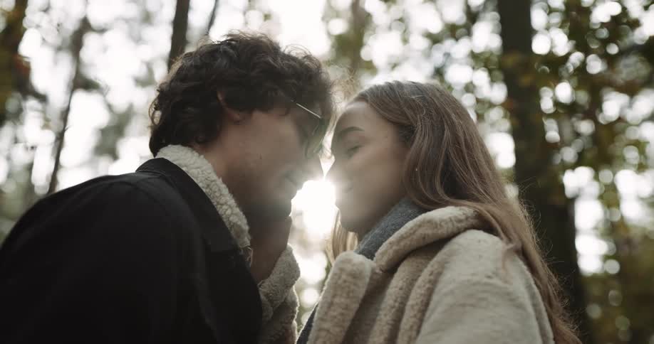 Man with glasses black jacket and backpack touches woman with brown hair beige coat and gray scarf in the autumn forest